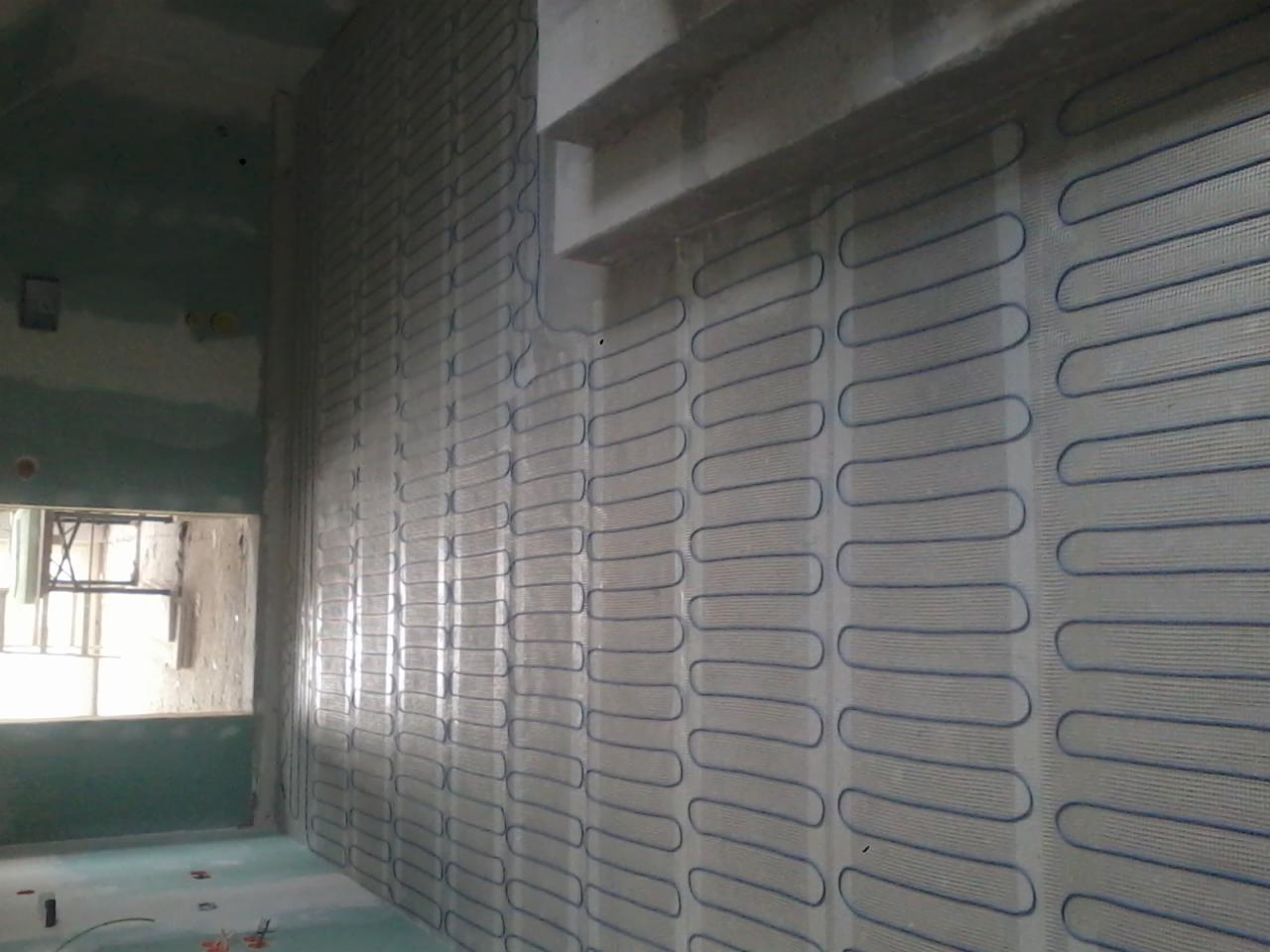 Ściana w trakcie montażu ogrzewania podłogowego ściennego, widoczne maty grzewcze z niebieskim przewodem układane pionowo na ścianie w surowym stanie, w tle okno.