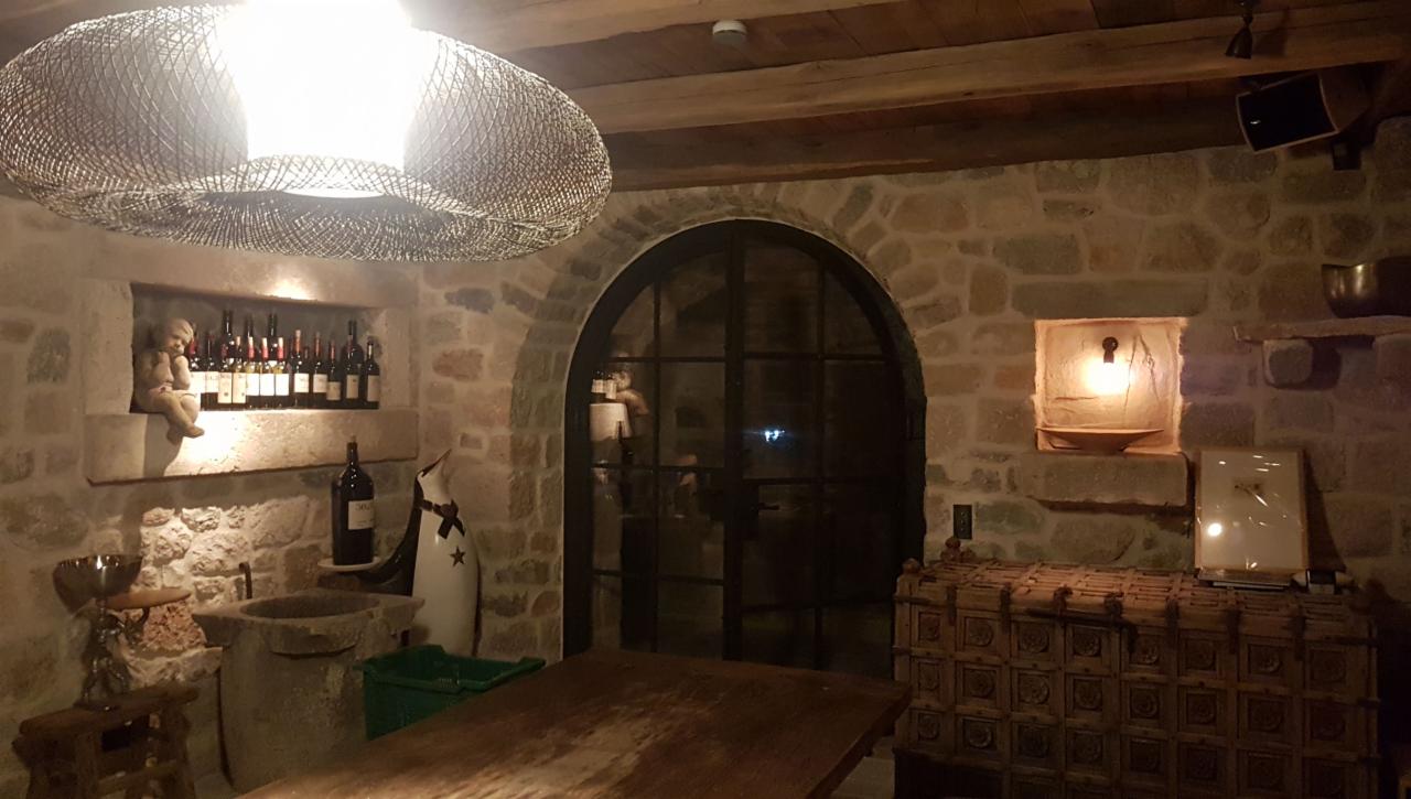 Rustykalna piwnica z kamiennymi ścianami, oświetlona wiszącą lampą z abażurem z plecionki, z dekoracyjnymi niszami, w których znajdują się butelki wina, figurka aniołka i świeca, oraz drewniany...