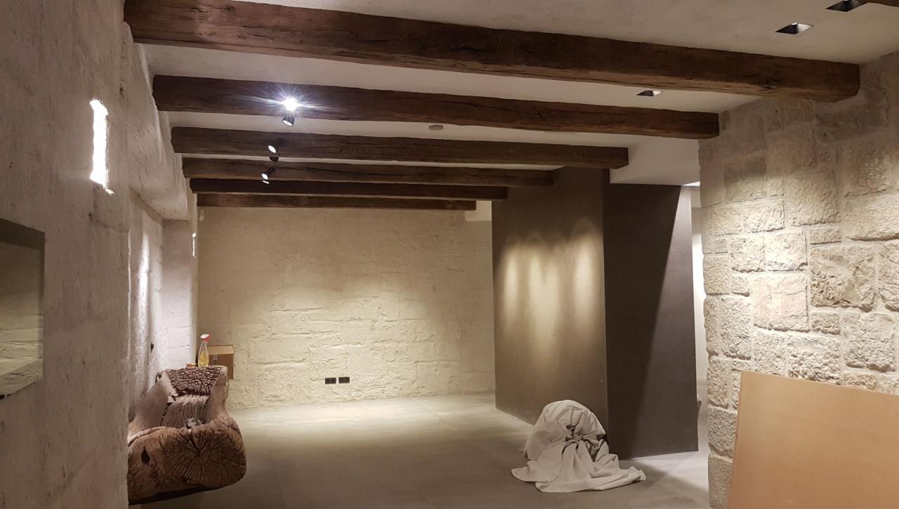 Surowe wnętrze z beżowymi ścianami o nierównej fakturze, drewnianymi belkami stropowymi i punktowym oświetleniem, w tle widoczna kolumna w kolorze ciemnego beżu, na podłodze leży biała tkanina.