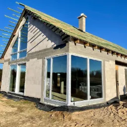 Surowy stan budynku z dużymi oknami z białymi ramami, widoczne etapy budowy dachu i komina, teren wokół budynku w stanie naturalnym, piasek i ziemia.