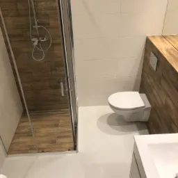 Nowoczesna łazienka z kabiną prysznicową wyłożoną płytkami imitującymi drewno, białą ceramiką i drewnopodobną zabudową.