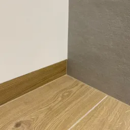 Szczeg&oacute;łowe ujęcie rogu pomieszczenia z drewnopodobną podłogą, listwą przypodłogową i szarą ścianą wykończoną płytkami imitującymi beton.