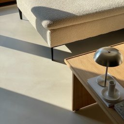 Kadap Sp z o.o. - Minimalistyczne wnętrze z beżową sofą, drewnianym stolikiem i lampą w metalicznym odcieniu, oświetlone naturalnym światłem, akcentujące gładką, jasną podłogę.