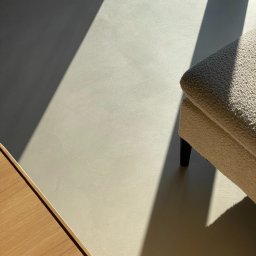 Kadap Sp z o.o. - Jasna posadzka, prawdopodobnie z mikrocementu, oświetlona mocnym światłem słonecznym, obok fragment beżowego fotela i drewnianej szafki. Minimalistyczne wnętrze.