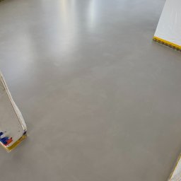 Kadap Sp z o.o. - Gładka, szara posadzka z mikrocementu w nowym wnętrzu. Ściany zabezpieczone folią i taśmą. Widoczne narożniki pomieszczenia i fragment okna w górnym rogu.