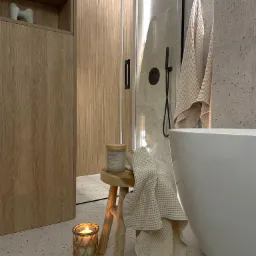 Stylowa łazienka z kabiną prysznicową typu walk-in, drewnianą zabudową i wolnostojącą wanną. Na stołeczku świeca i ręcznik w odcieniach beżu, tworząc przytulną atmosferę.