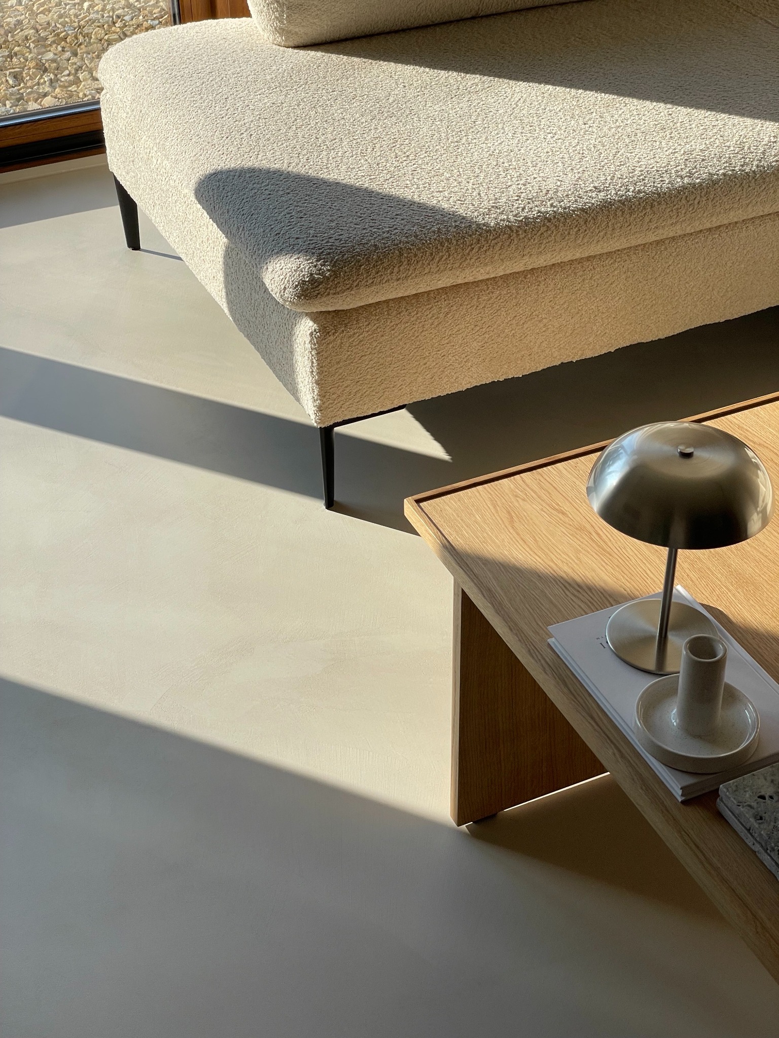 Minimalistyczne wnętrze z beżową sofą, drewnianym stolikiem i lampą w metalicznym odcieniu, oświetlone naturalnym światłem, akcentujące gładką, jasną podłogę.