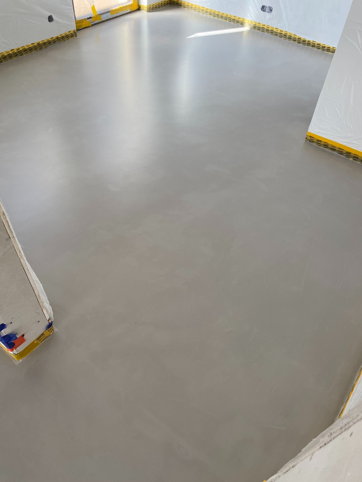 Gładka, szara posadzka z mikrocementu w nowym wnętrzu. Ściany zabezpieczone folią i taśmą. Widoczne narożniki pomieszczenia i fragment okna w górnym rogu.