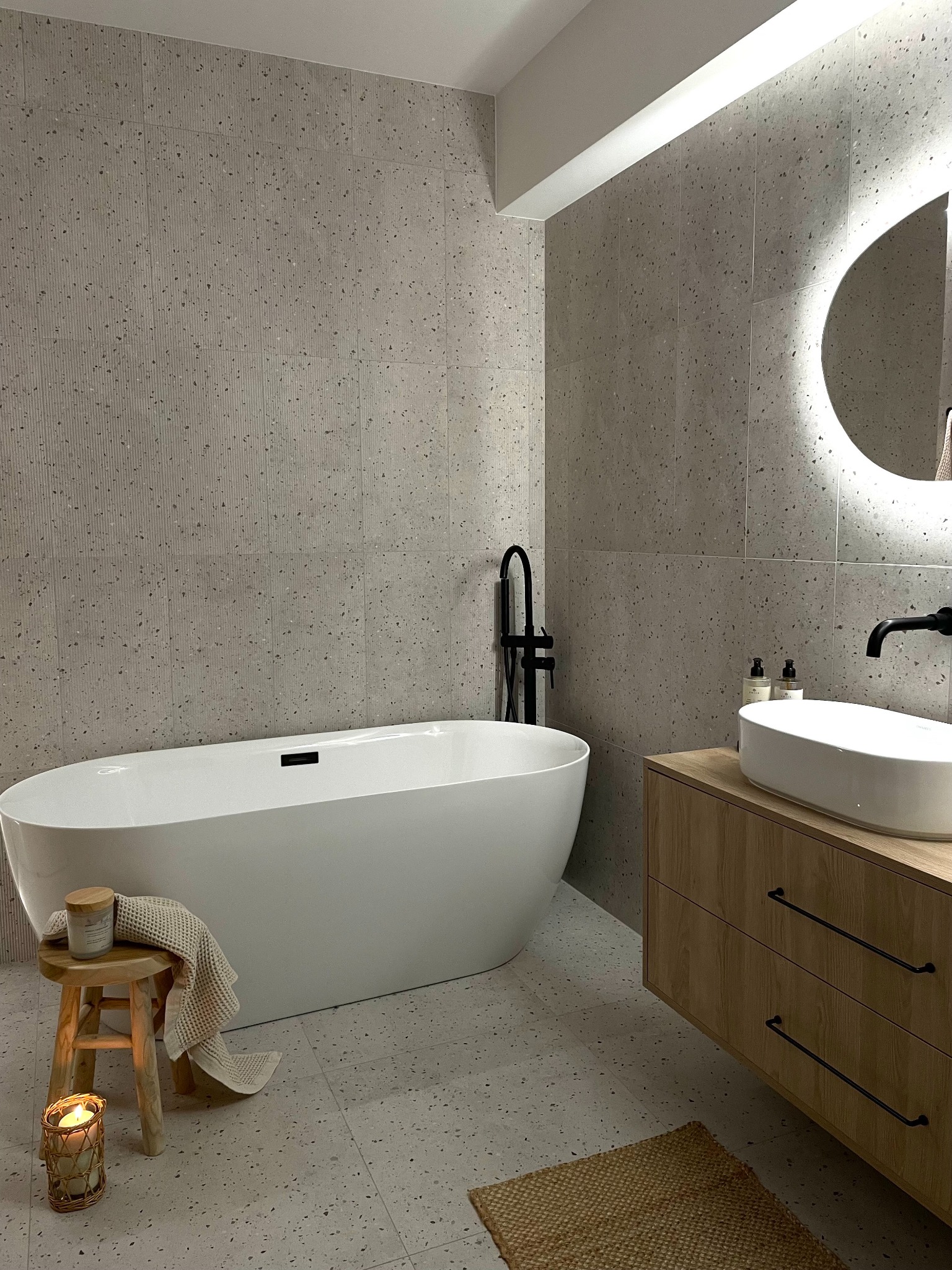 Stylowa łazienka z wanną wolnostojącą, szarą mozaiką na ścianach i podłodze, drewnianą szafką z umywalką oraz okrągłym lustrem z podświetleniem LED.