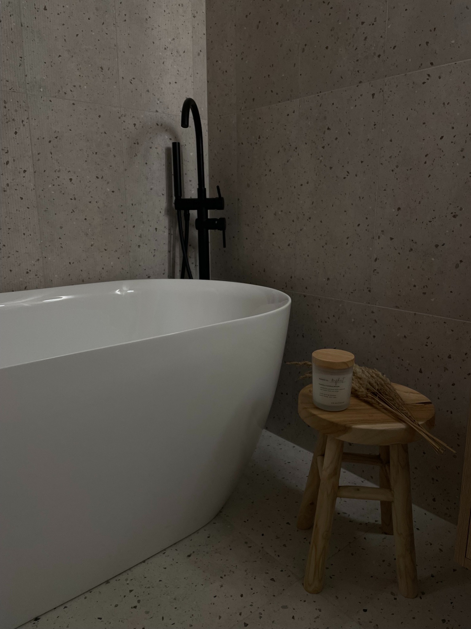 Minimalistyczna łazienka: owalna wanna, czarna bateria, terazzo na ścianach i podłodze. Drewniany stołek z dekoracyjną świecą i suszem.