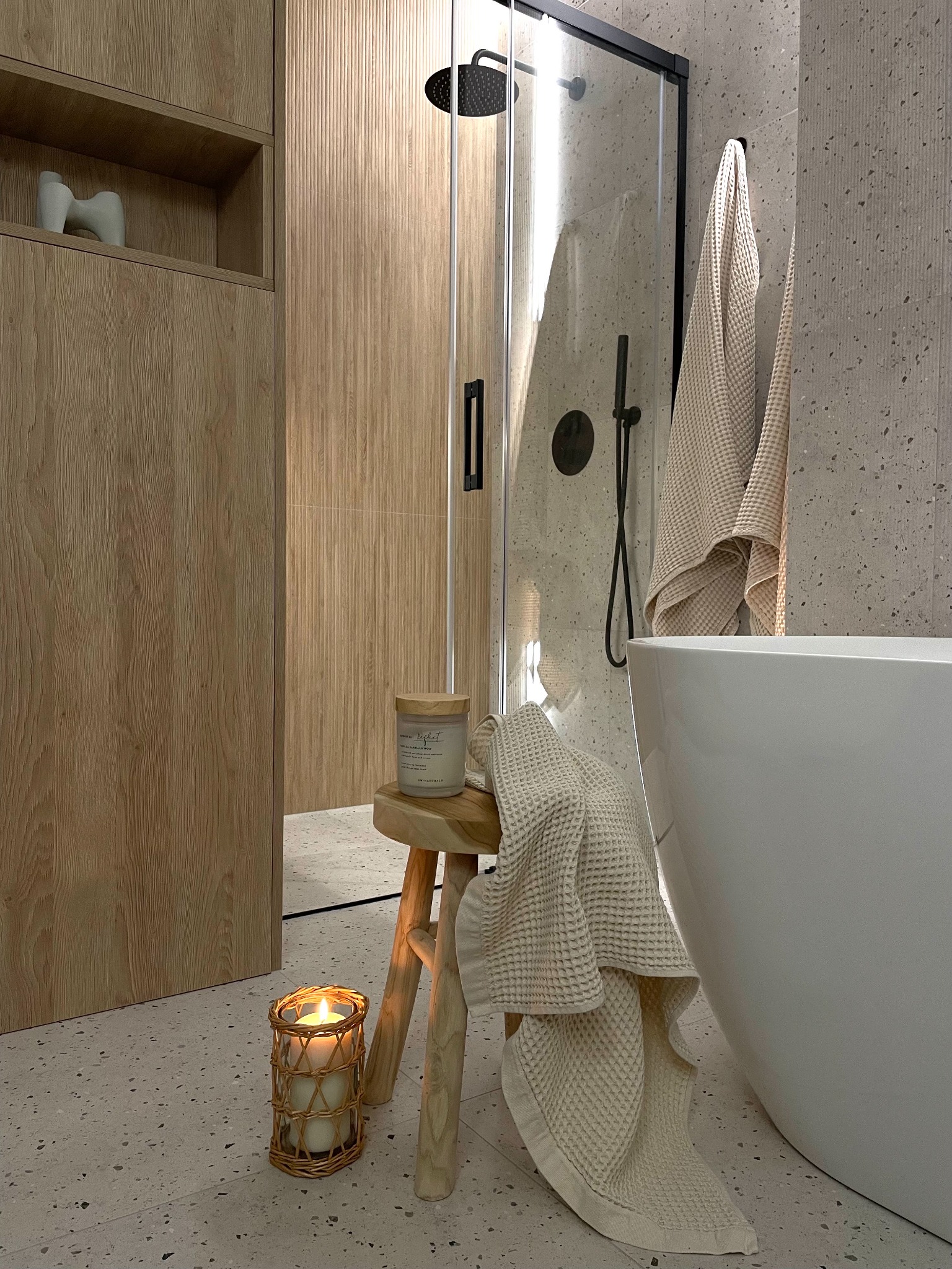 Stylowa łazienka z kabiną prysznicową typu walk-in, drewnianą zabudową i wolnostojącą wanną. Na stołeczku świeca i ręcznik w odcieniach beżu, tworząc przytulną atmosferę.