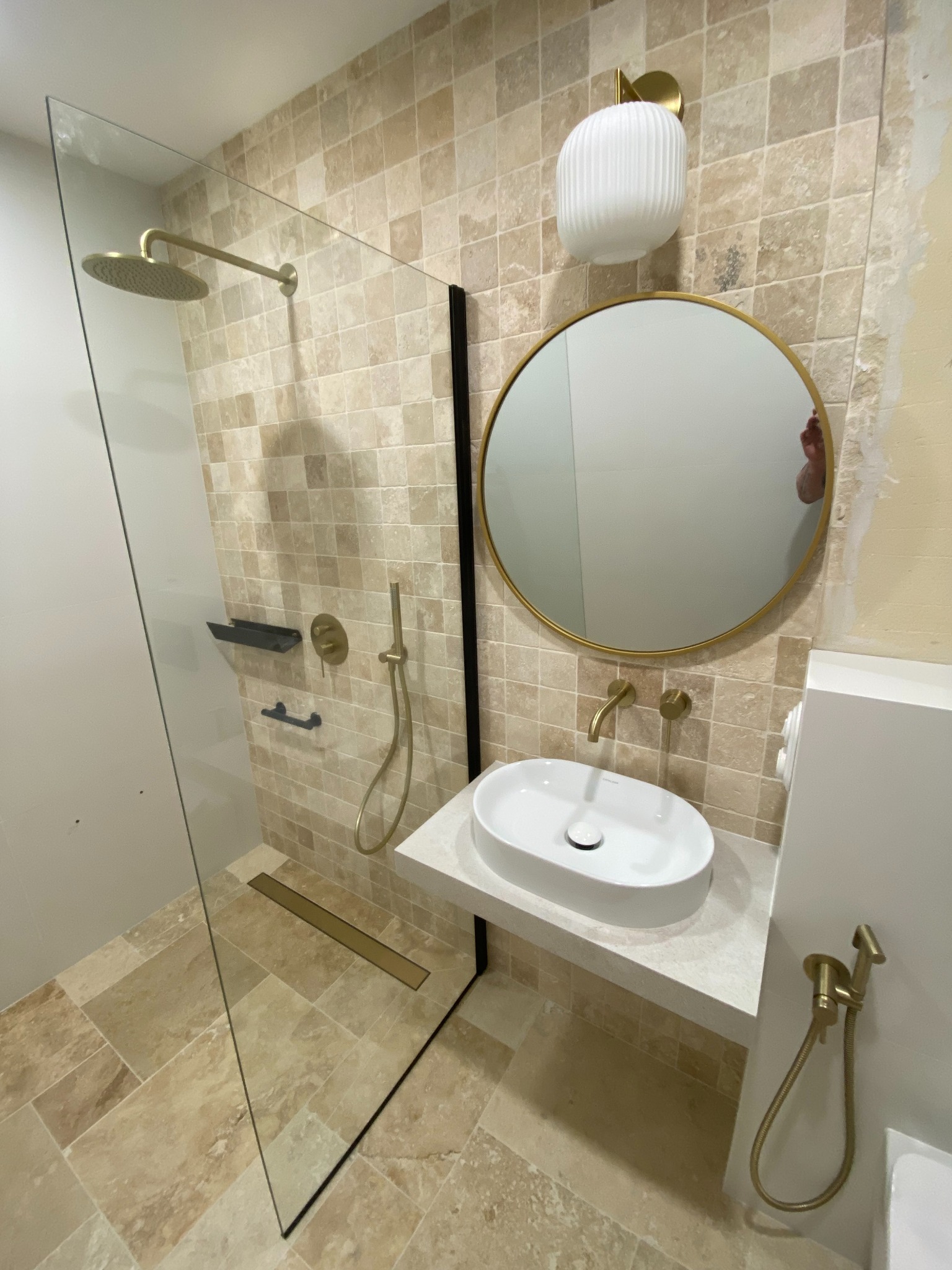 Elegancka łazienka z kabiną prysznicową typu walk-in, umywalką nablatową i złotymi akcentami armatury na tle trawertynowych płytek. Minimalistyczny design z okrągłym lustrem.