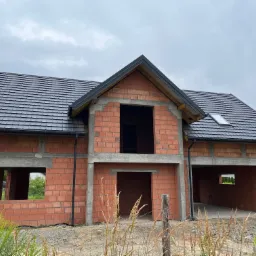 Surowy stan domu jednorodzinnego z czerwonej cegły w Warszawie, z czarną dachówką i widocznymi elementami konstrukcji żelbetowej, na tle pochmurnego nieba.