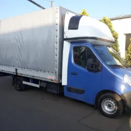 Niebieski samochód dostawczy Renault Master z plandeką, zaparkowany na asfaltowej drodze, widok z boku, z tłem zielonych krzewów i białego budynku.