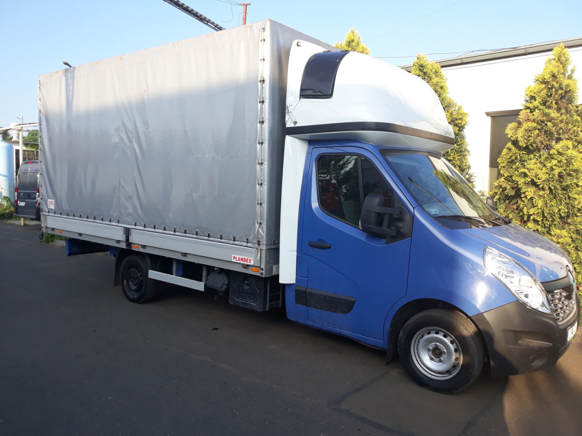 Niebieski samochód dostawczy Renault Master z plandeką, zaparkowany na asfaltowej drodze, widok z boku, z tłem zielonych krzewów i białego budynku.