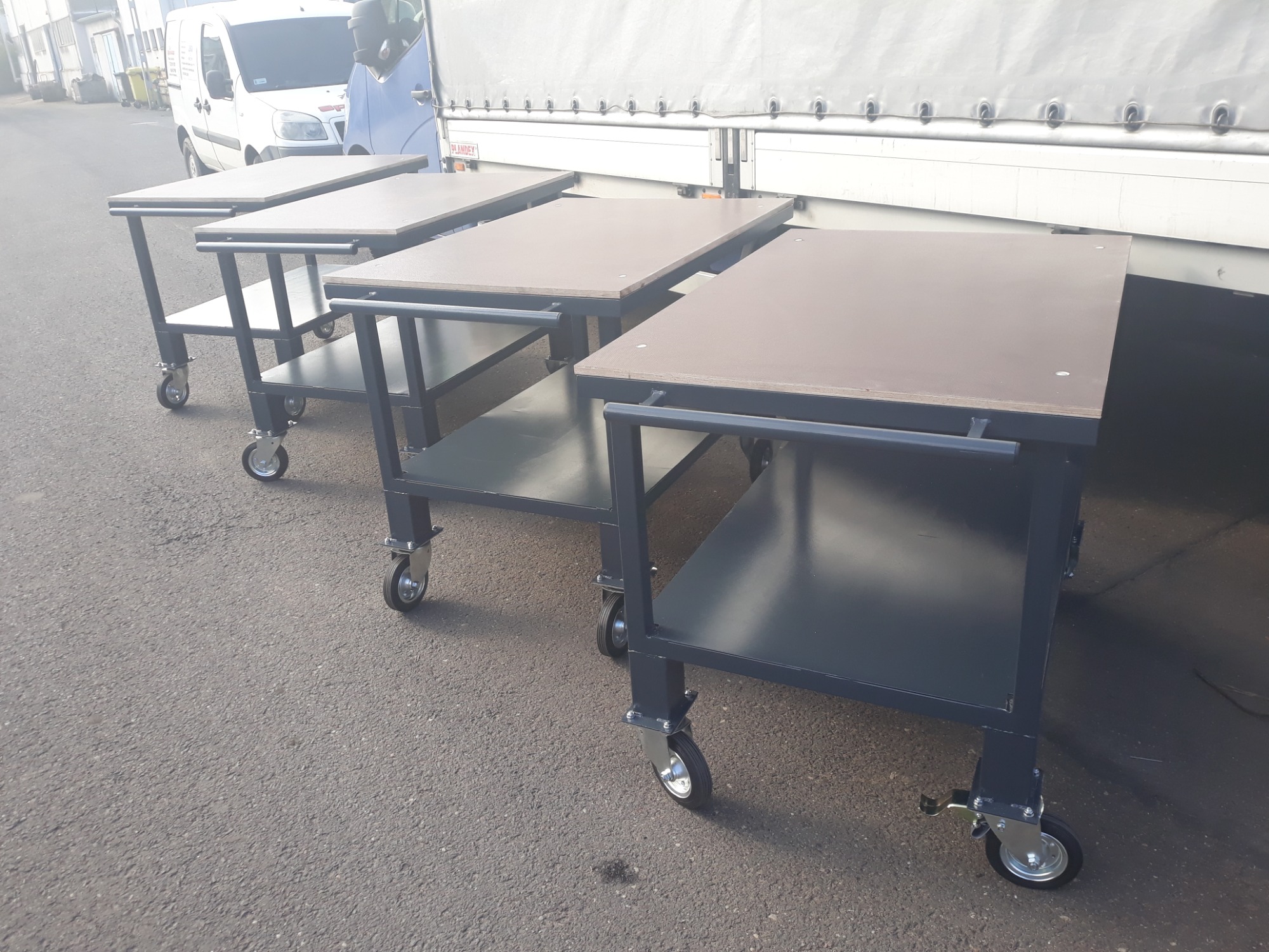 Cztery stoły transportowe produkcyjne z metalową konstrukcją i kółkami, ustawione na zewnątrz na tle białej plandeki samochodu dostawczego.