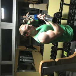 Mężczyzna w zielonym podkoszulku podnosi sztangę w siłowni, widoczne odzwierciedlenie w lustrze i stojak z ciężarami w tle.