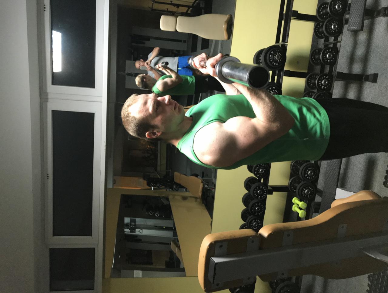 Mężczyzna w zielonym podkoszulku podnosi sztangę w siłowni, widoczne odzwierciedlenie w lustrze i stojak z ciężarami w tle.