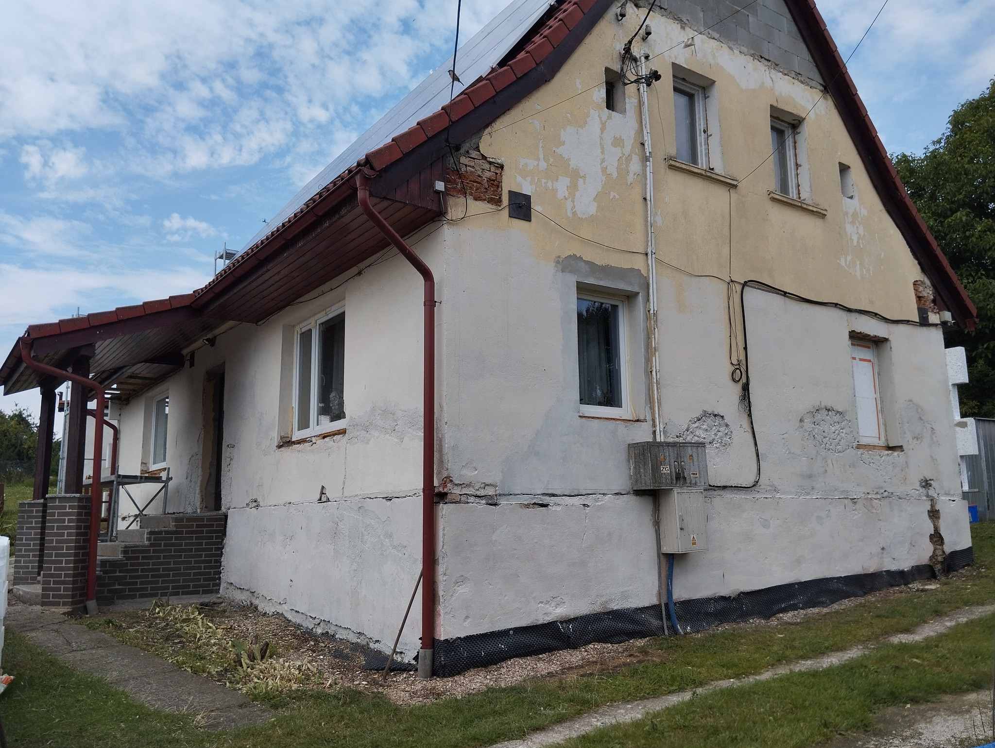 Dwupiętrowy dom z częściowo otynkowaną elewacją, widoczne ślady po uszkodzeniach i odpadającym tynku, panele słoneczne na dachu, czerwone rynny i podmurówka zabezpieczona folią fundamentową.