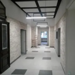 Wykończony korytarz biurowy z ceglanymi ścianami, czarnym sufitem podwieszanym z oświetleniem LED, szarą podłogą z kwadratowymi wstawkami i drewnianą drabiną w tle.