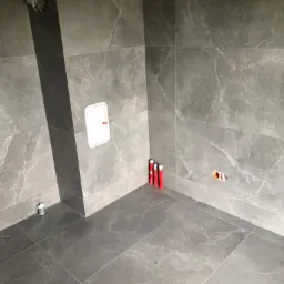 Wnętrze łazienki w trakcie prac w Pierzchnicy, widoczne szare płytki imitujące marmur na ścianach i podłodze, otwór rewizyjny, wystające rury i otwory instalacyjne.