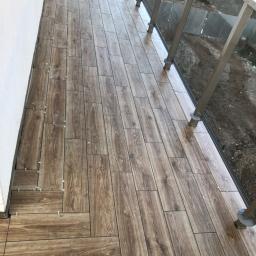 PRO-BERT Norbert Wojtyna - Balkon w trakcie wykańczania, brązowe płytki imitujące drewno ułożone w jodełkę, widoczne krzyżyki dystansowe, szklana balustrada.