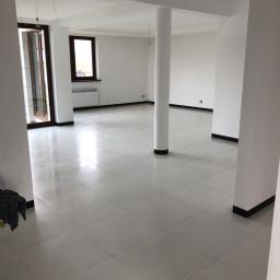 PRO-BERT Norbert Wojtyna - Jasne, przestronne wnętrze z białymi ścianami, kolumną i podłogą wyłożoną błyszczącymi, jasnymi płytkami. Widoczne okna z brązowymi ramami i grzejnik. Minimalistyczne wykończenie.