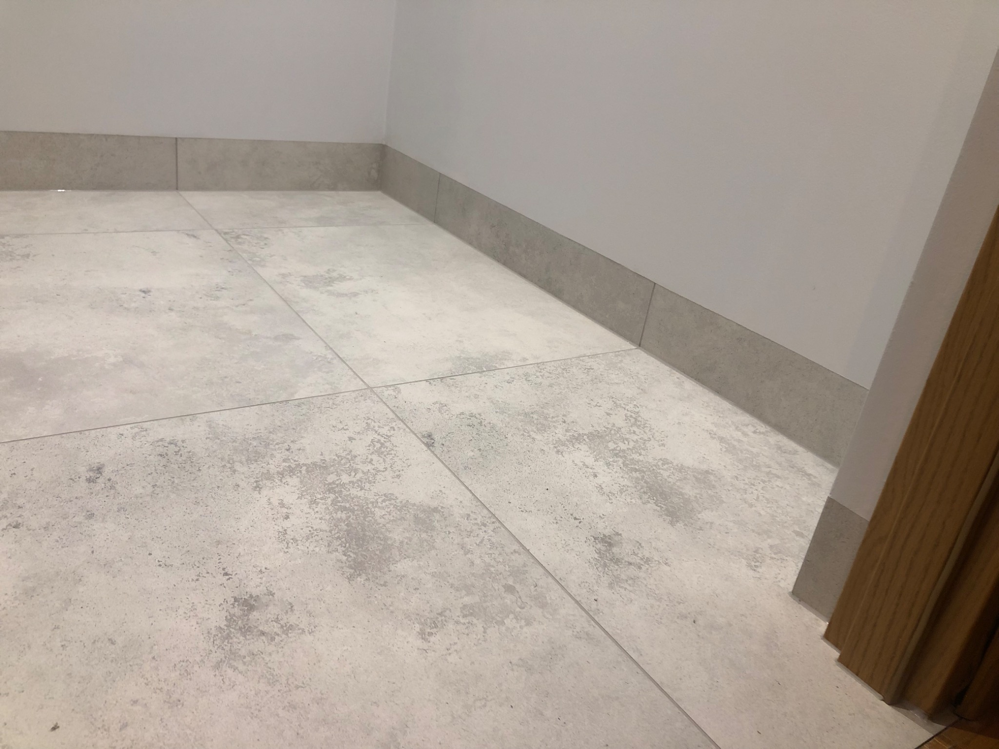 Jasnoszara, matowa podłoga z płytek imitujących beton, z dopasowaną listwą przypodłogową, przy białej ścianie i drewnianej futrynie drzwiowej.