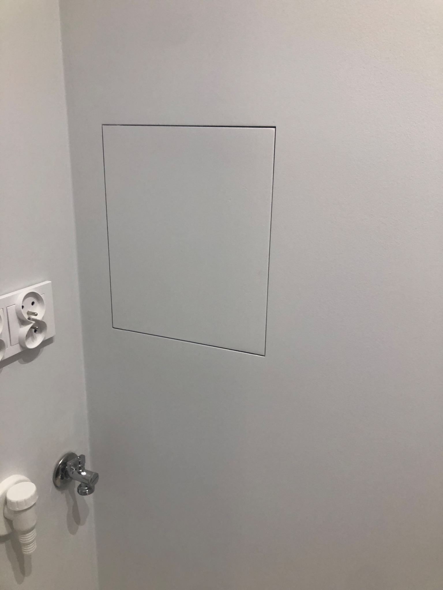 Biała ściana w łazience z ukrytym włazem rewizyjnym, gniazdkiem elektrycznym i zaworem wody.