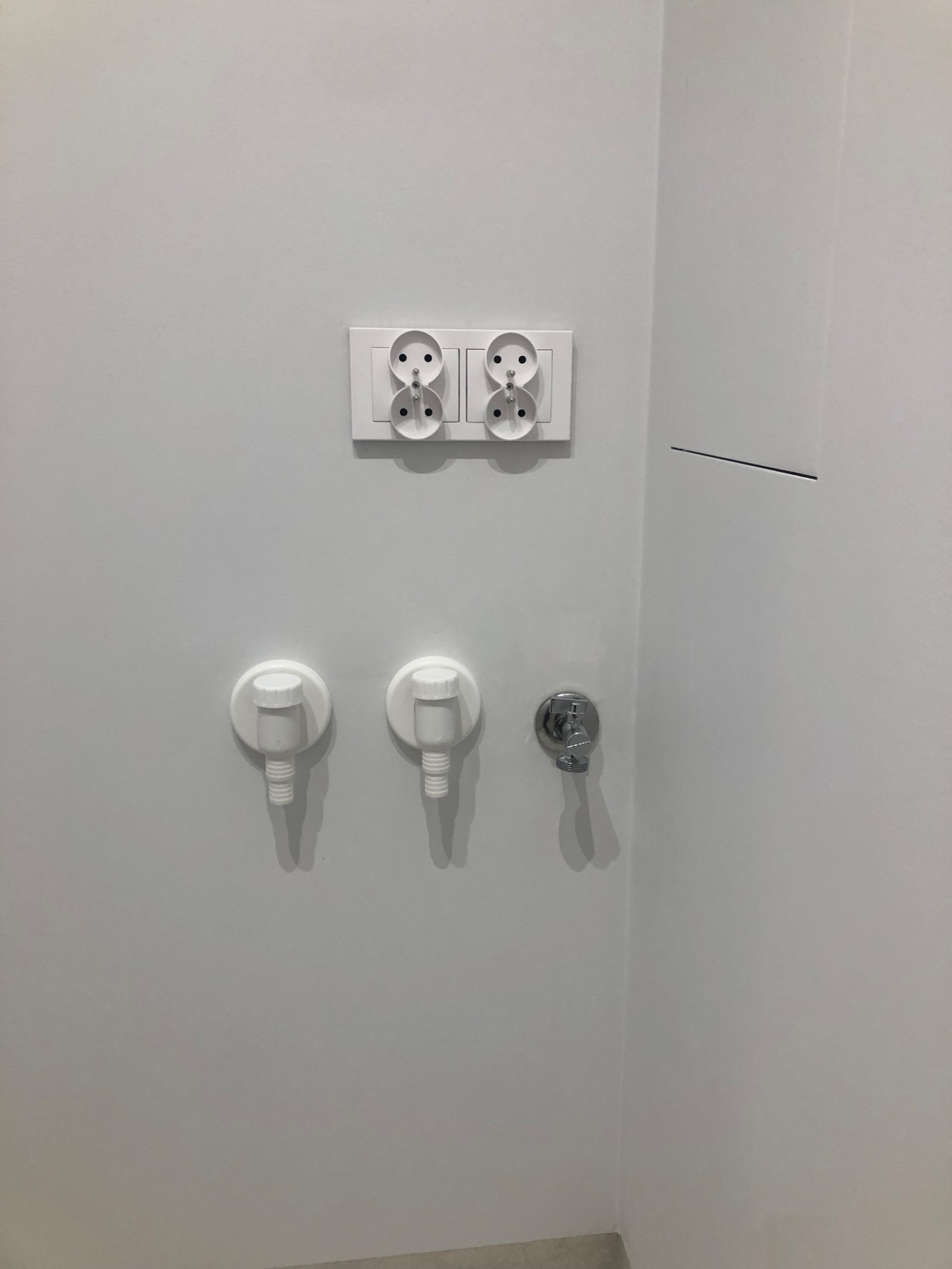 Biała ściana łazienki z podwójnym gniazdkiem elektrycznym, dwoma zaworami do podłączenia pralki i chromowanym zaworem wody