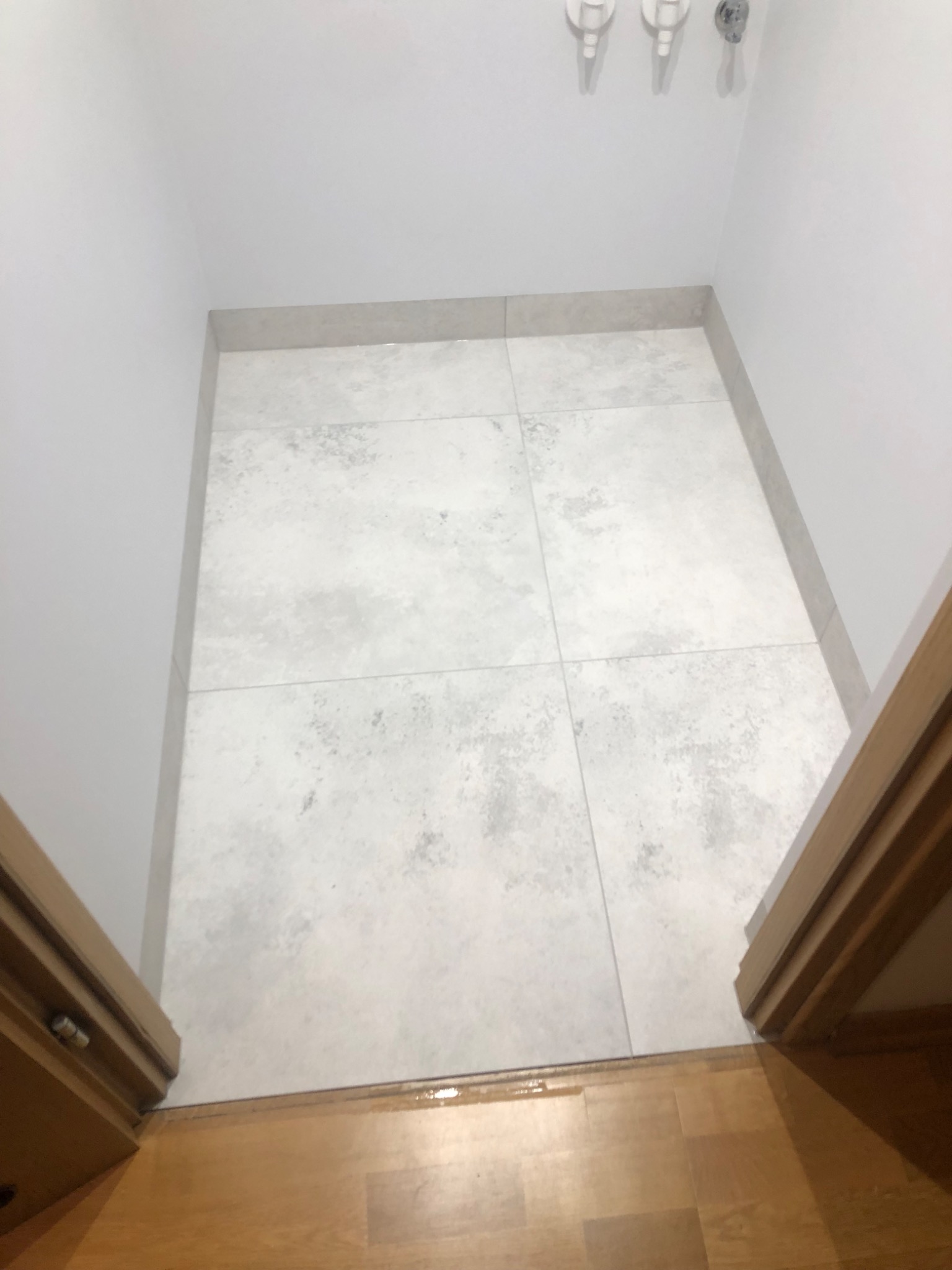 Jasnoszara, kwadratowa podłoga wyłożona płytkami w małym pomieszczeniu, widok z góry na wejście z drewnianą ościeżnicą i parkietem.