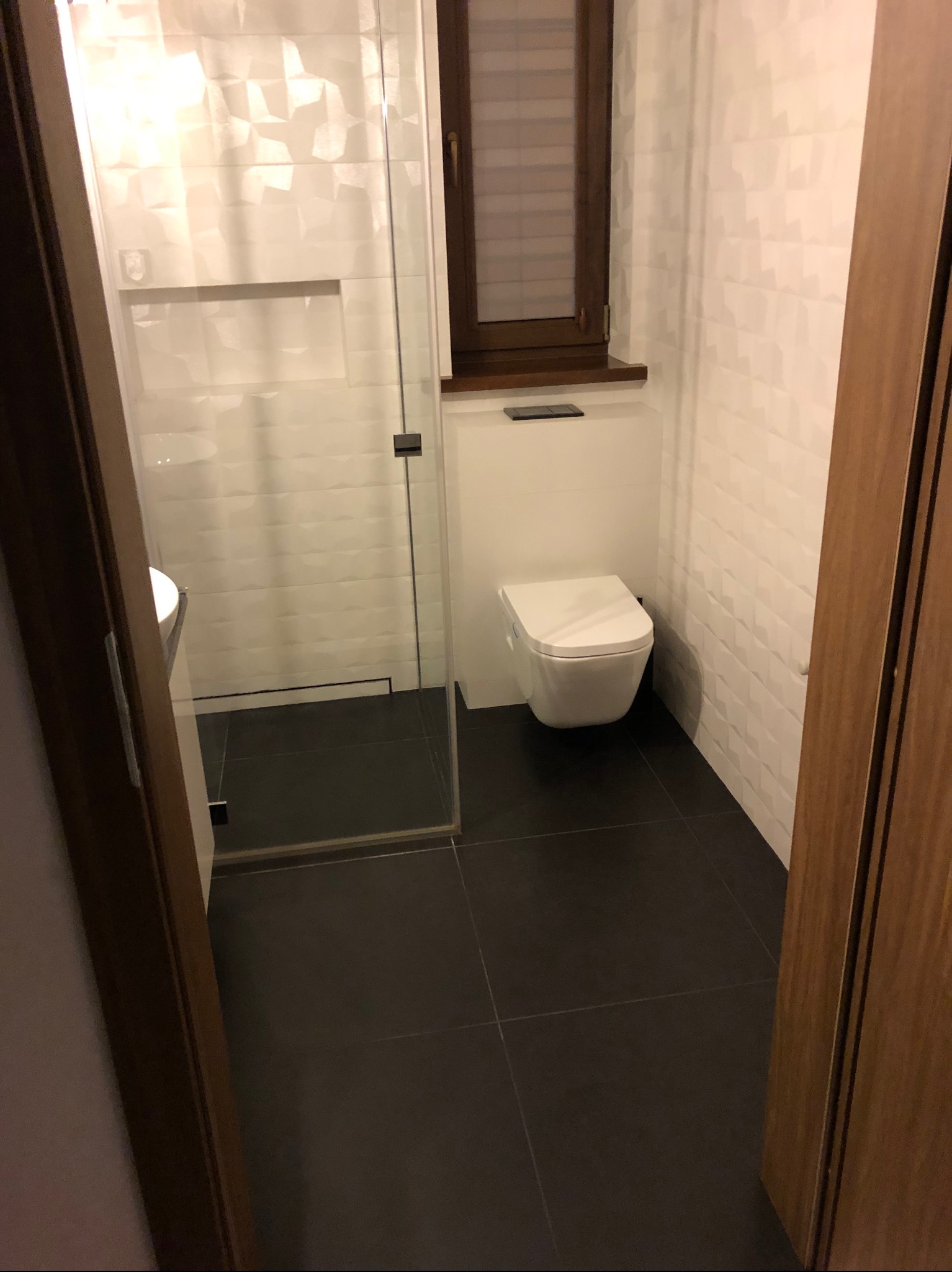 Nowoczesna łazienka z czarną podłogą, białą, trójwymiarową glazurą ścienną, podwieszaną toaletą i kabiną prysznicową ze szklanymi drzwiami.