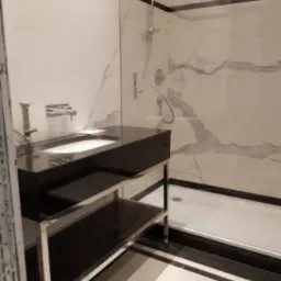 Nowoczesna łazienka z marmurowymi płytkami, czarną armaturą i szklaną kabiną prysznicową, widok na umywalkę z czarnym blatem i chromowanym stelażem.