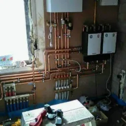 Rozbudowana instalacja centralnego ogrzewania z miedzianych rur, widoczne dwa białe piece grzewcze, rozdzielacze, liczne zawory i złączki, fragment ściany z cegły oraz prowizoryczny warsztat na tle...