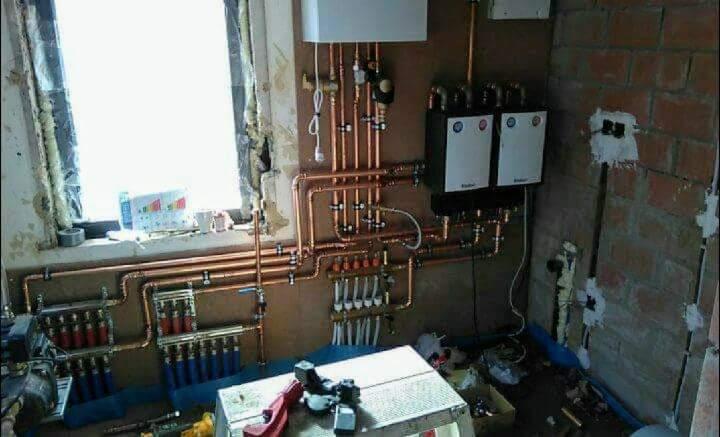 Rozbudowana instalacja centralnego ogrzewania z miedzianych rur, widoczne dwa białe piece grzewcze, rozdzielacze, liczne zawory i złączki, fragment ściany z cegły oraz prowizoryczny warsztat na tle...