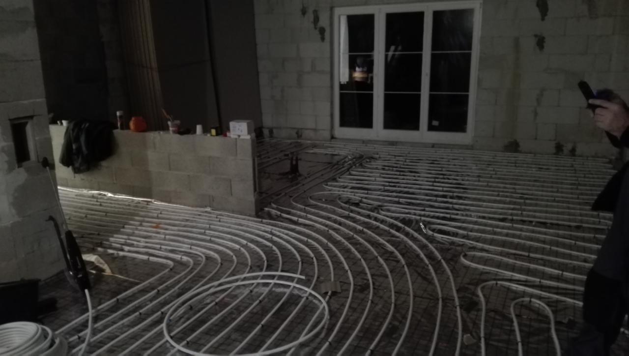 Rozłożone rury ogrzewania podłogowego na siatce w surowym wnętrzu, widoczne białe rury i fragment okna.