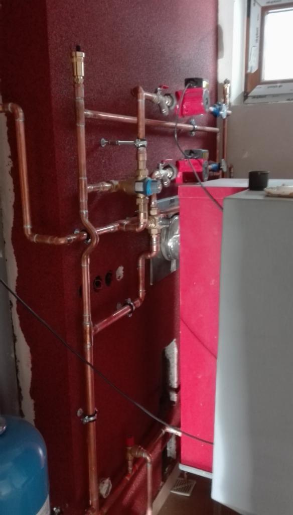 Świeżo zamontowana instalacja centralnego ogrzewania z miedzianymi rurami, czerwonymi pompami obiegowymi i zbiornikiem wyrównawczym, widoczne ślady po pracach na czerwonej ścianie.