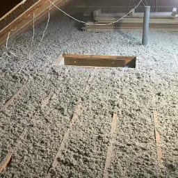 Poddasze z widoczną warstwą izolacji z wełny mineralnej, drewnianą konstrukcją krokwi i wystającymi przewodami elektrycznymi oraz kominem wentylacyjnym z PCV.