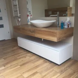 Nowoczesna łazienka z drewnianym blatem pod umywalkę nablatową i białymi szafkami podwieszanymi, jasne płytki na podłodze i ścianach, grzejnik drabinkowy.