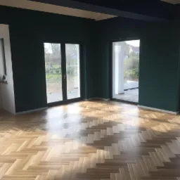 Wnętrze z nową, jasną parkietową podłogą ułożoną we wzór jodełki klasycznej, świeżo pomalowane ściany na ciemnozielono, duże okna balkonowe z widokiem na ogród.