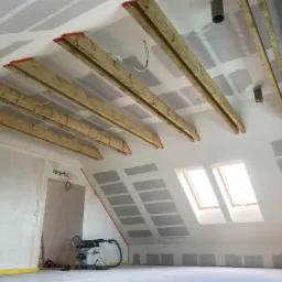 Poddasze w trakcie adaptacji: widoczne drewniane belki konstrukcyjne, częściowo zamontowane płyty gipsowo-kartonowe na skosach, narzędzia budowlane na podłodze, dwa okna dachowe wpuszczające jasne...