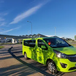 Jaskrawozielony bus Opel Vivaro z pomarańczową kamizelką odblaskową na desce rozdzielczej, zaparkowany na tle lotniska i błękitnego nieba z lekkimi chmurami.