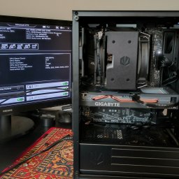 Zbigniew Derus - Wnętrze czarnej obudowy komputera stacjonarnego z widoczną kartą graficzną Gigabyte i chłodzeniem procesora, obok monitor z ekranem BIOS, prezentacja konfiguracji.