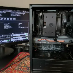 Wnętrze czarnej obudowy komputera stacjonarnego z widoczną kartą graficzną Gigabyte i chłodzeniem procesora, obok monitor z ekranem BIOS, prezentacja konfiguracji.