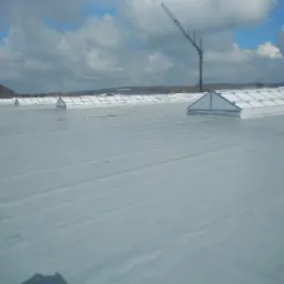 Widok z góry na płaski dach pokryty jasnoszarą folią elastyczną, z widocznymi świetlikami dachowymi w kształcie piramid i żurawiem budowlanym w tle.