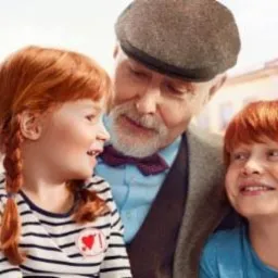 Uśmiechnięty dziadek w kaszkiecie i muszce z wnuczkami o rudych włosach na tle rynku miejskiego.