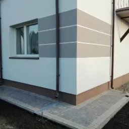 Dwukondygnacyjny budynek z ocieploną elewacją w odcieniach bieli i szarości, z brązowym cokołem i nową kostką brukową wokół, widoczne rury spustowe i okno z białymi ramami.