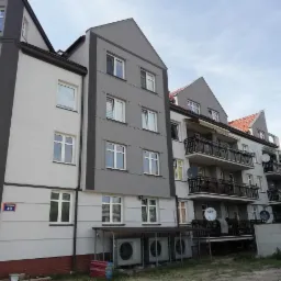 Odnowiony budynek wielorodzinny z szarą i białą elewacją, widoczne balkony z czarnymi balustradami i anteny satelitarne, numer budynku 49 na ścianie, oraz klimatyzatory na parterze.