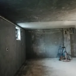 Surowe wnętrze w trakcie remontu: szare ściany z widoczną fakturą tynku, małe okno, instalacje elektryczne i hydrauliczne, podnośnik na trójnogu na środku pomieszczenia.