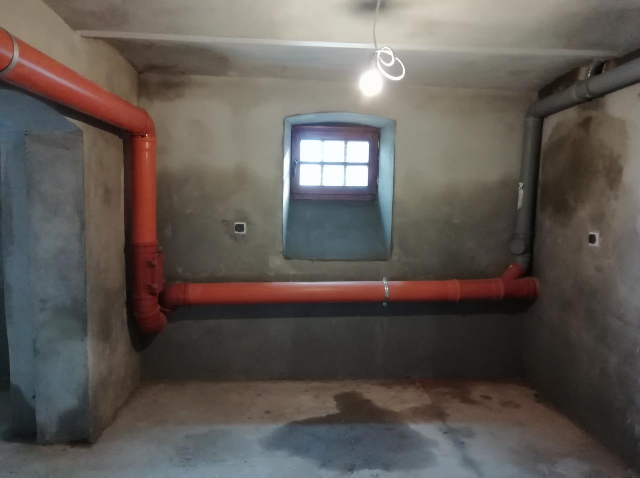 Pomieszczenie w trakcie remontu z widocznymi rurami kanalizacyjnymi w kolorze pomarańczowym i szarym, małe okno z drewnianą ramą oraz surowymi ścianami i podłogą z plamami.